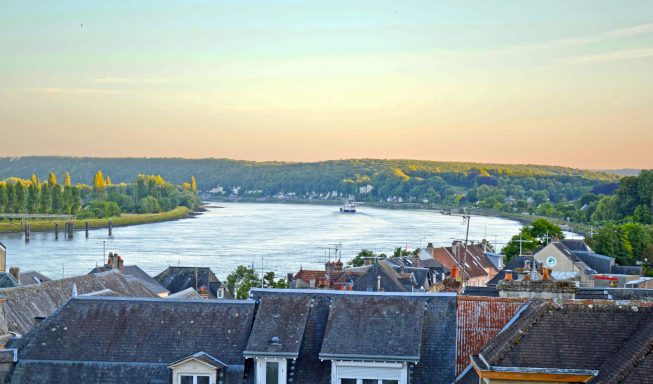 Blick auf einen Fluss umgeben von grünen Hügeln und Dächern im Sonnenuntergang.